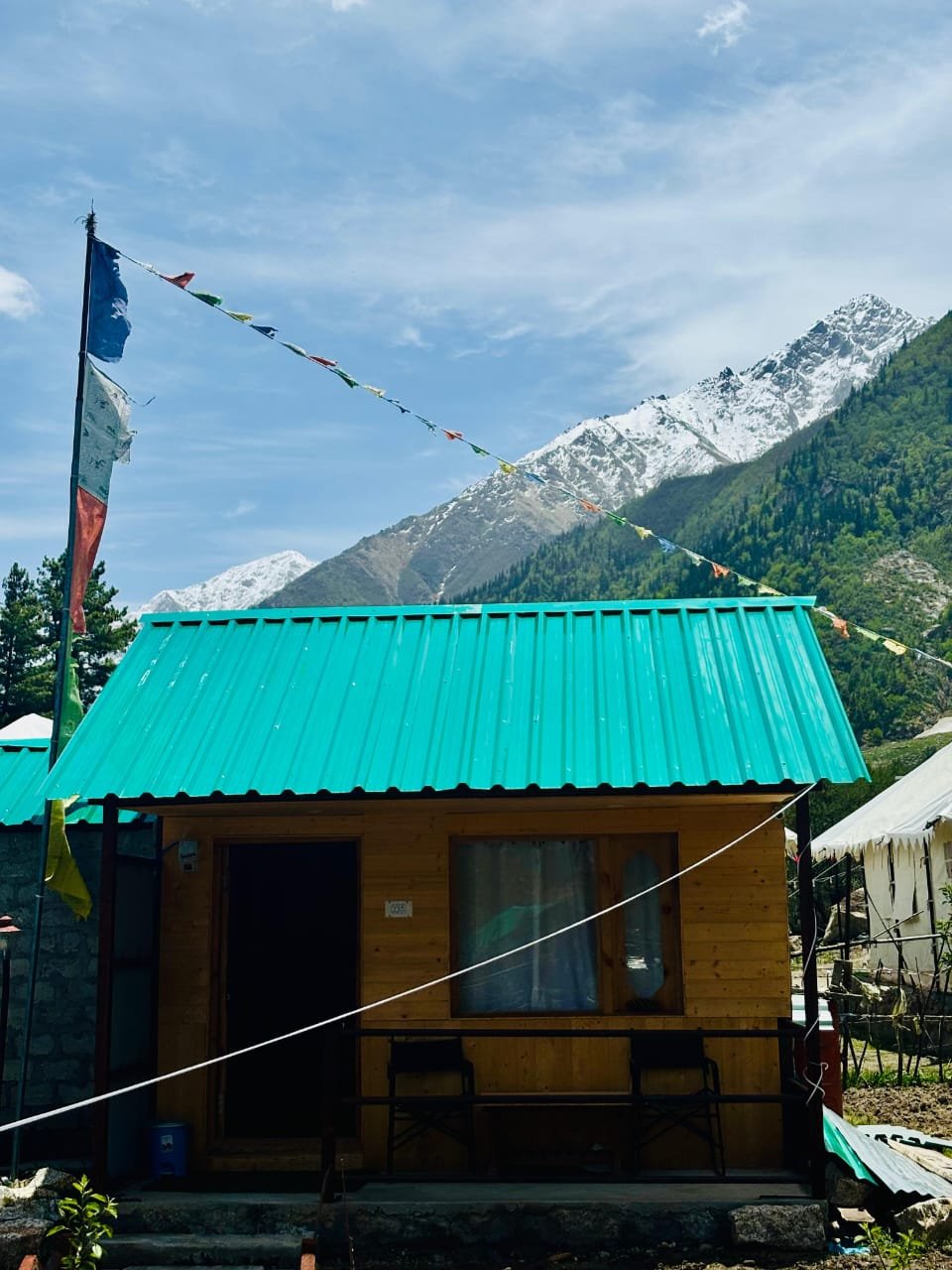 Pine cottage with teal roof, prayer flags, and snow peaks rising behind