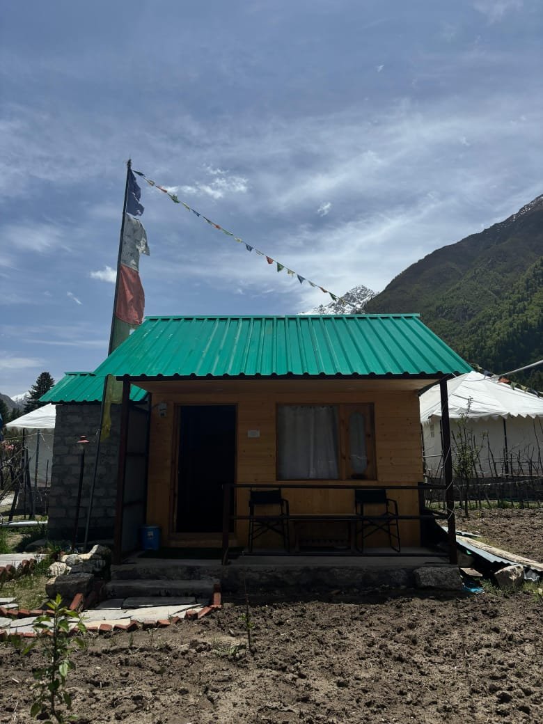Pine cottage exterior with teal roof, prayer flags, chairs on verandah, Himalayan peaks behind